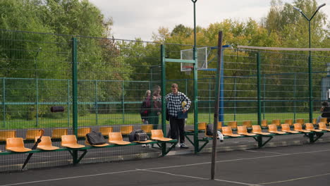 volleyball players with sports bags enter outdoor court, preparing for warm-up run with coach, they wear casual athletic attire, carrying sports equipment, court has orange seats and metal fence