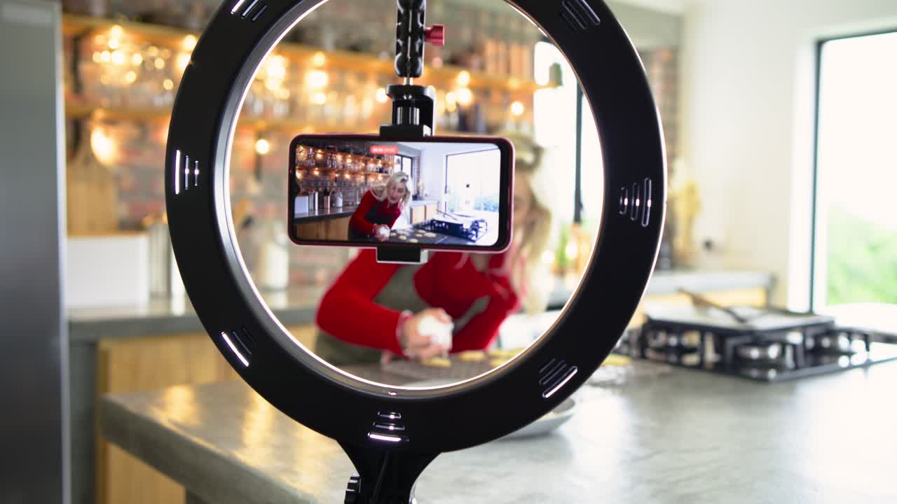 Seeing smartphone in ring light, adult female piping heart-shaped cookie icing demo in kitchen