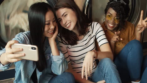 three pretty mixed races friendly girls at washing machines in washhouse smiling to smartphone camera, taking selfire photo