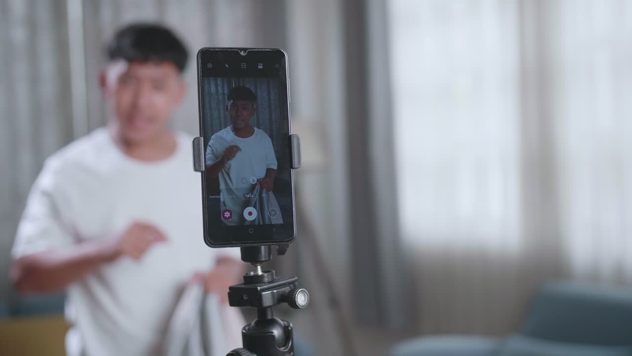 Lo schermo dello smartphone di un ragazzo asiatico mostra un trucco di sparizione sulla fotocamera del telefono cellulare mentre registra contenuti video per i social network in una stanza accogliente.