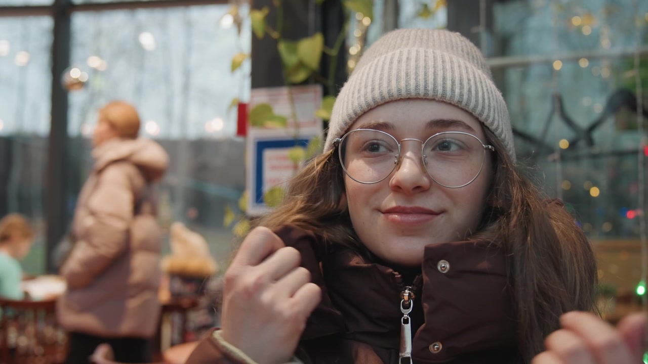 Donna bianca in un accogliente mercato che si allaccia la giacca, giovane professionista che sistema il colletto, con berretto di lana e occhiali rotondi, un sorriso sottile, luci festive e acquirenti sullo sfondo, un'atmosfera calma e sicura.