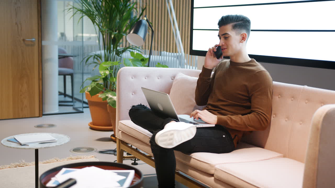 Young white businessman using phone working on a laptop computer sitting on a sofa in a casual office