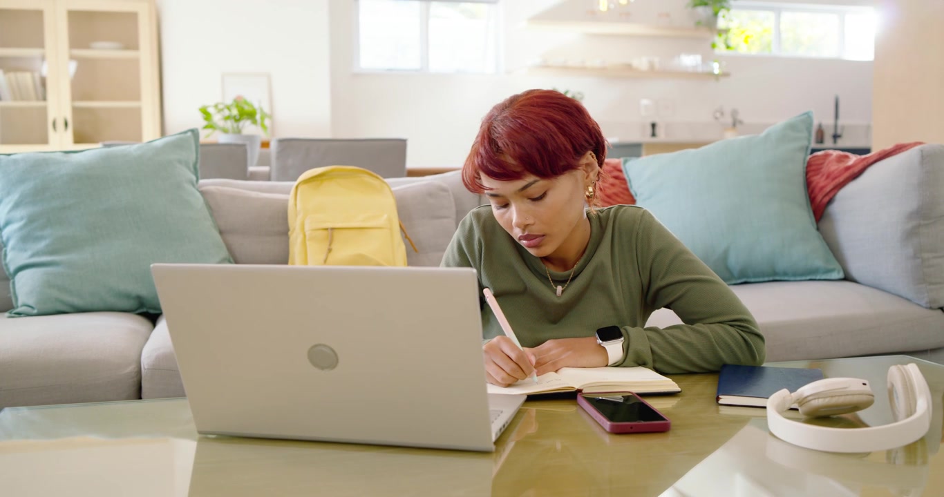 African American woman watching laptop, pausing, writing notes at living room coffee table studying