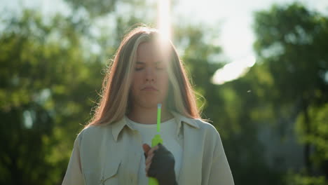 blonde-haired girl standing thoughtfully, holding green air pump with focused expression as sunlight creates radiant effect on her head, set against background of lush trees