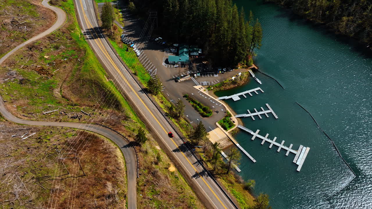 Red automobile approaching the boat station near the beautiful pine tree forest. Drone footage following the car.