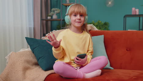 Child-girl-kid-with-smartphone-in-headphones-dancing-singing-listening-music-at-home-alone-on-sofa