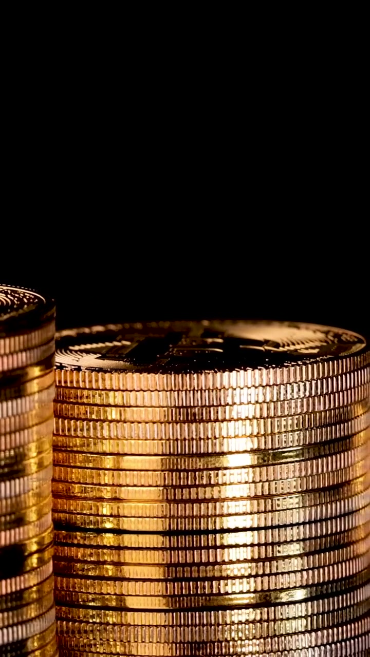 Macro vertical shot of shiny gold coin stacks rotating against a dark, reflective background.
