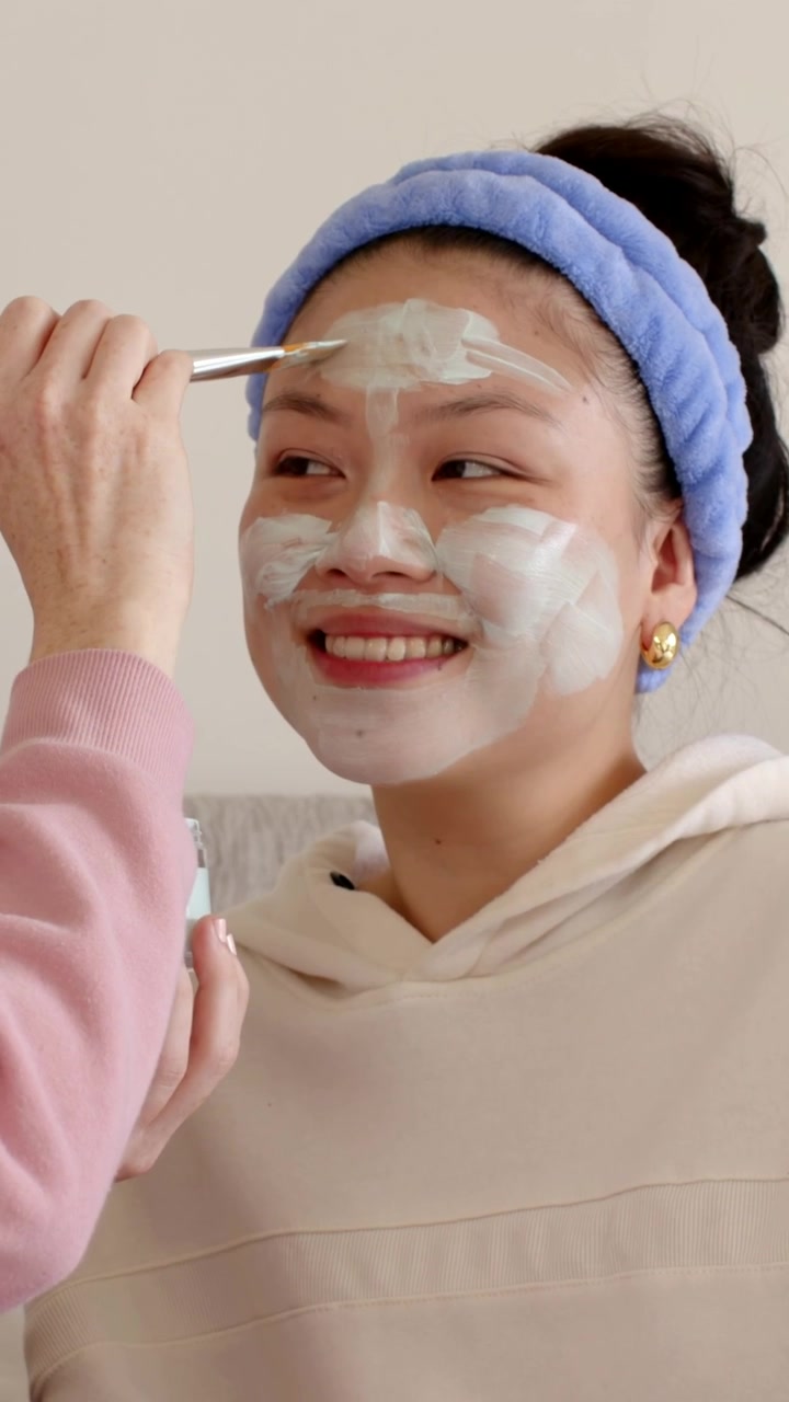 Video vertikal: Kuas muncul, asisten mengoleskan masker pada wanita di rumah yang mengenakan bando, dengan toples.