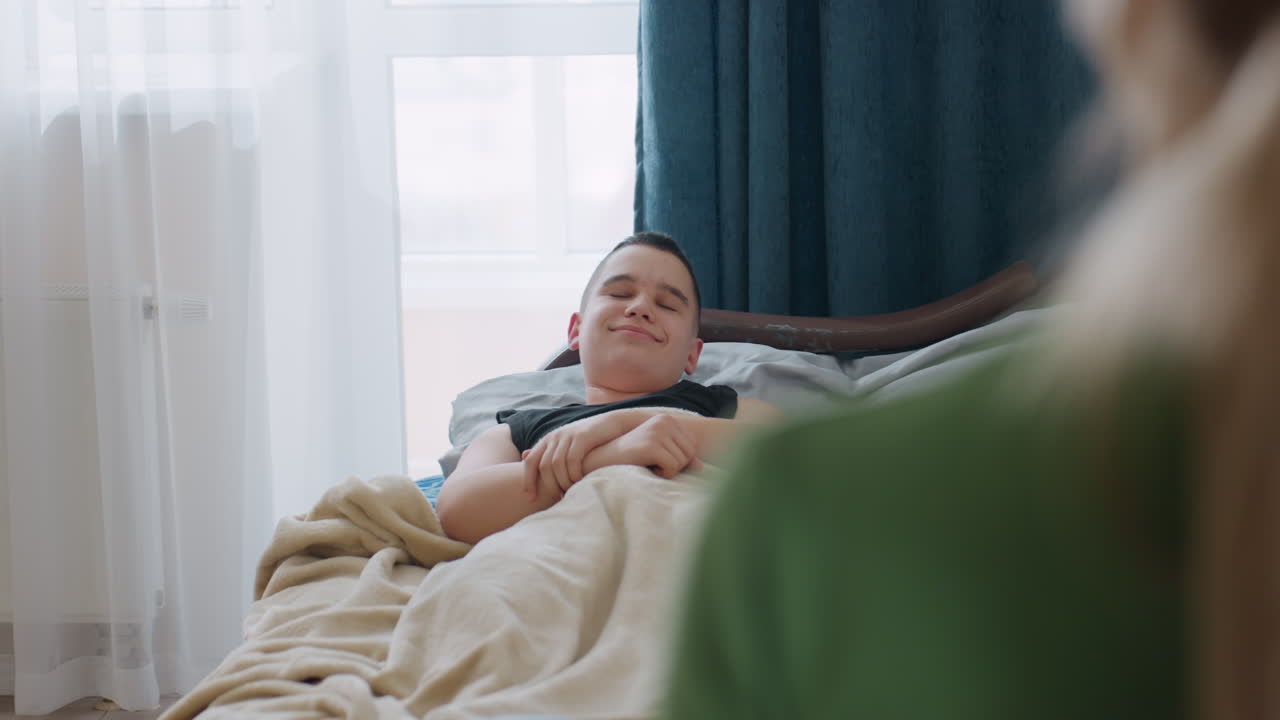 Little boy wakes up stretching under blanket in cozy bedroom filled with daylight as mother comes in to check on him while he smiles warmly creating tender family bonding