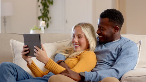 diverse couple relaxing on couch, smiling while watching tablet at home