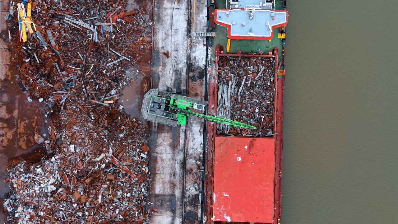 Veduta aerea dall'alto di una gru che carica rottami metallici su una nave. Movimentatore di materiali idraulico verde che sposta rifiuti metallici riciclati nella stiva di una nave da carico in porto.