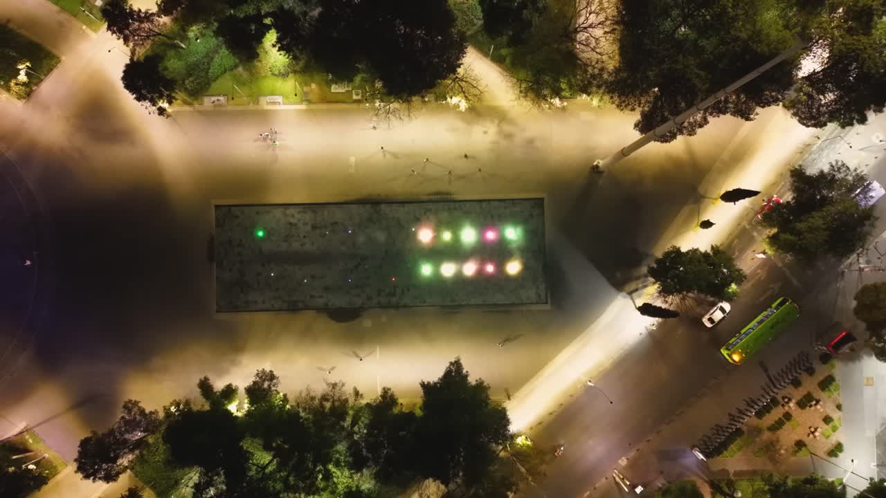 Filmagem noturna de drone de uma fonte iluminada com cores mutáveis em uma praça mexicana.