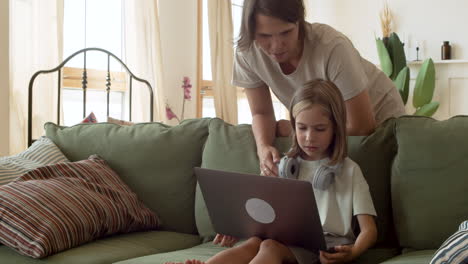 una niña rubia haciendo su tarea con la computadora portátil y su madre detrás de ella ayudando con la tarea