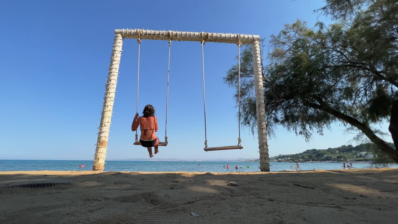 vista trasera de ángulo bajo de una mujer adulta en vacaciones de verano disfruta columpiándose en un columpio de cuerda mirando el mar en calma