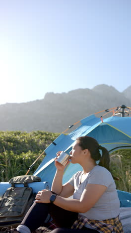 Vertical video: Camping in mountains, woman drinking from cup near tent, enjoying nature