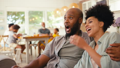 couple at home on sofa streaming show or movie to tv with multi-generation family in background