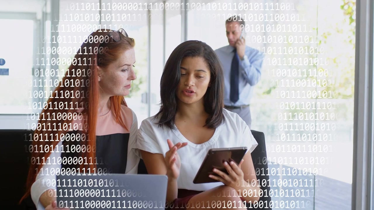 Two women sitting on office bench, right woman tapping tablet, showing tech data overlay, reviewing
