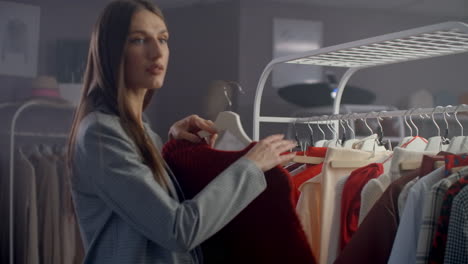 joven hermosa en una tienda de ropa buscando vestidos y suéteres en el mostrador. para tomar de una percha y para poner a sí misma sonriendo.