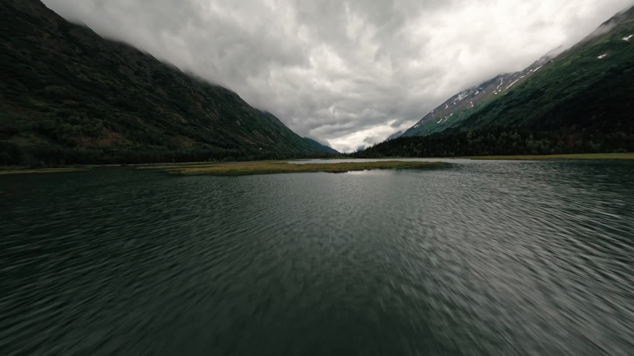 FPV aerial drone flying over Alaskan lake surrounded by mountains and lush flora on cloudy day