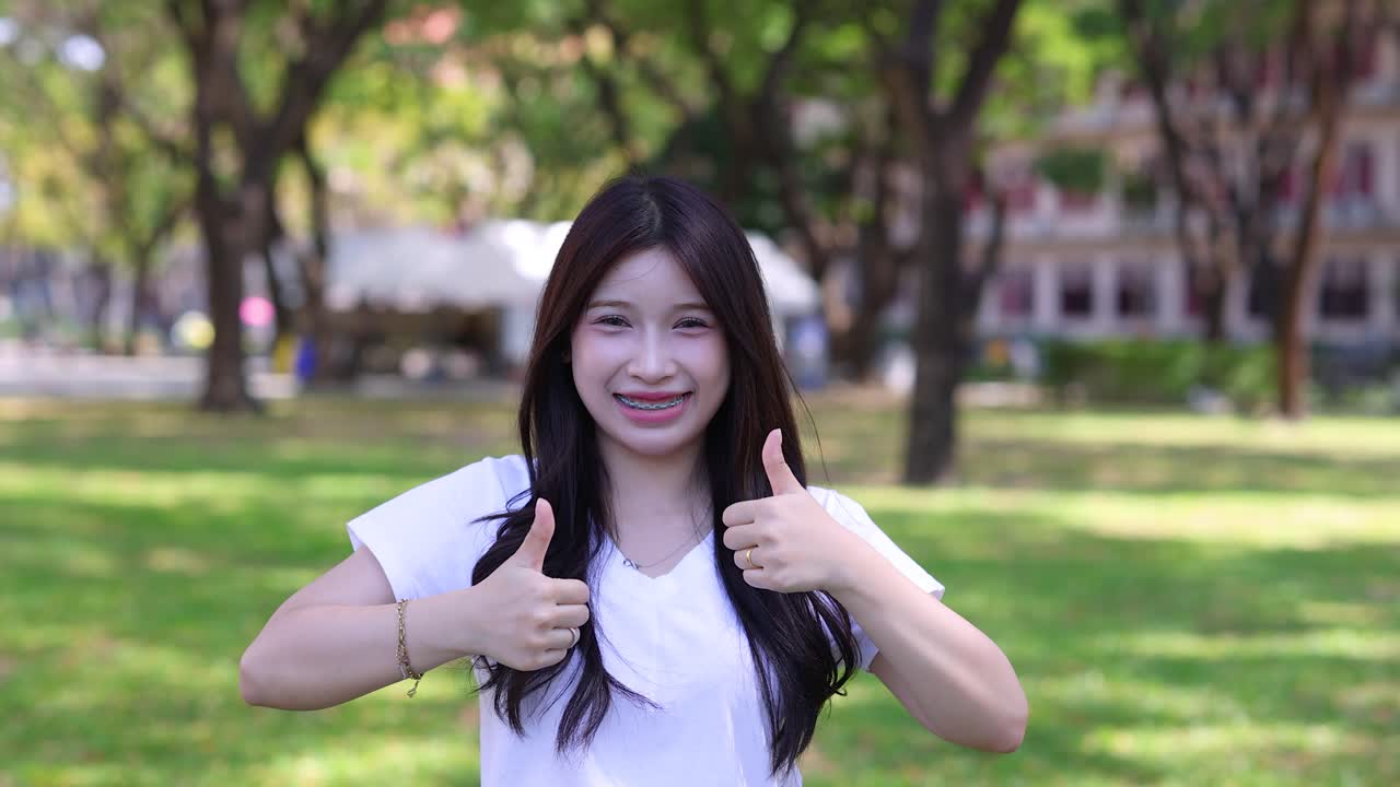 明るい屋外公園で、幸せそうなタイ人大学生が微笑んで親指を立てている。