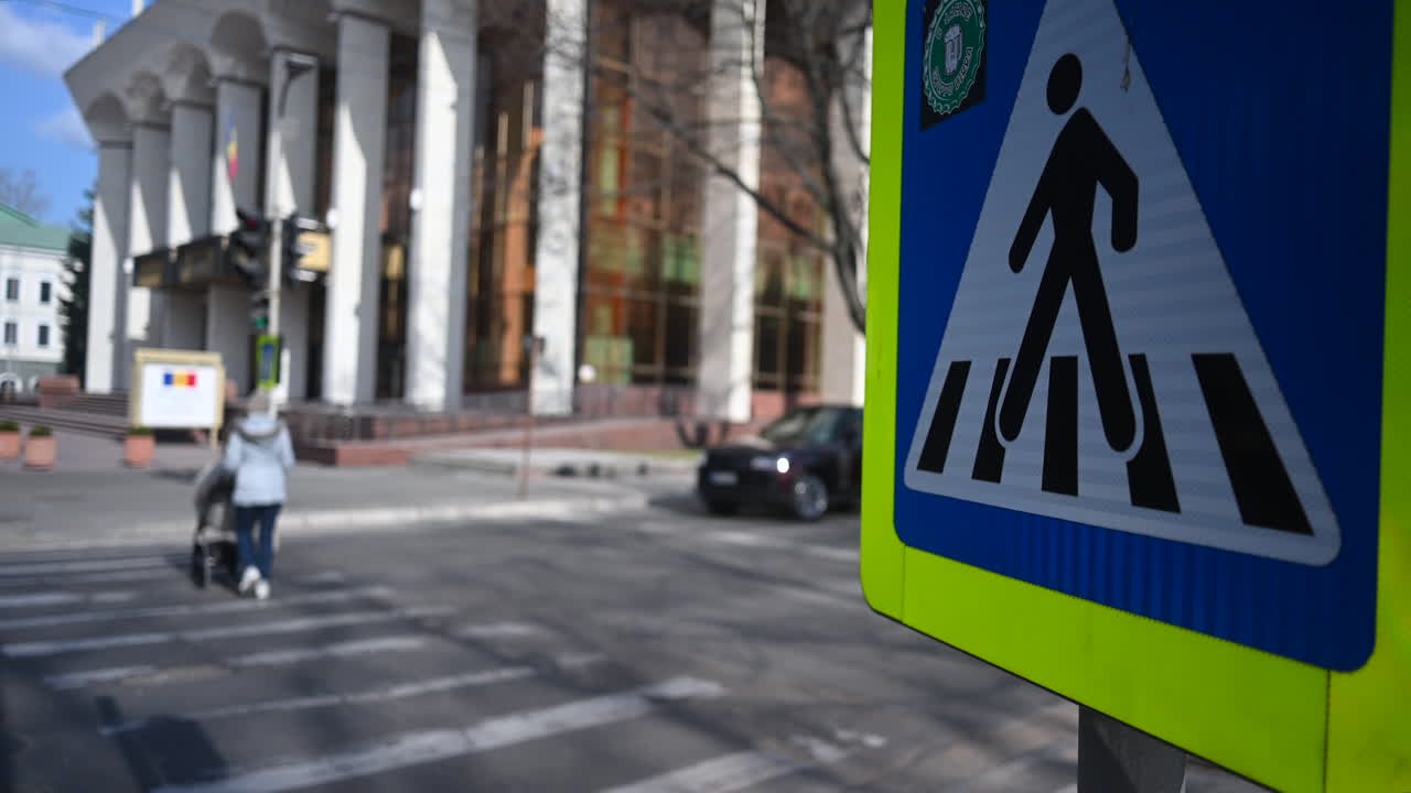 Chisinau, Moldavia - 18 marzo 2026: Segnale stradale di attraversamento pedonale a fuoco con una donna che spinge un passeggino sulle strisce pedonali in città.