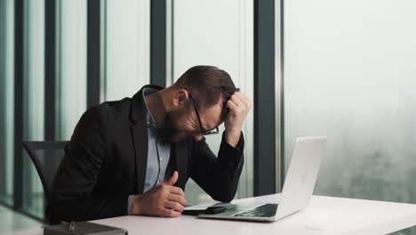 tired upset man takes off glasses while sitting at laptop in office near panoramic window