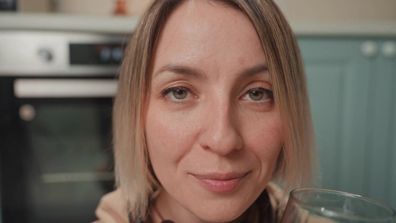 Donna bianca che beve acqua dopo lo yoga, primo piano di un bicchiere su una tavola di legno sul pavimento della cucina, studentessa di yoga che allunga la mano per prendere il bicchiere dal tappetino, coach del benessere che dimostra una routine di idratazione, creatrice di contenuti lifestyle che sorseggia.