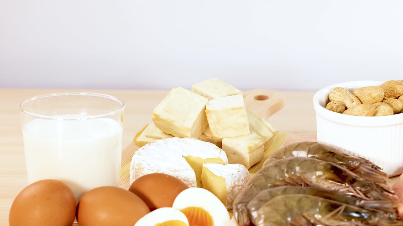 una variedad de alimentos ricos en proteínas, incluidos nueces, lácteos, huevos y mariscos, se exhiben en una superficie de madera con iluminación brillante