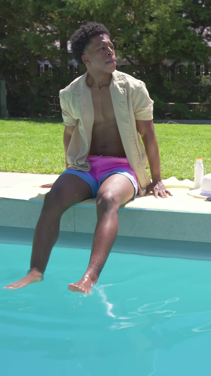 Vertical video: Walking man from lawn in pink trunks testing pool water sitting at deck near towel