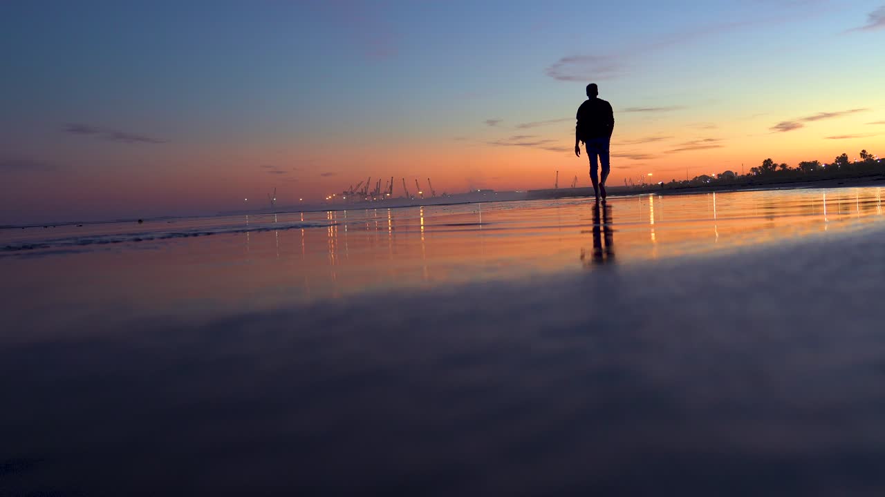 Man walks towards the horizon on beach at sunset, backlit with an orange sky and blue the beach inclined with the sand and the reflection of water on the slope