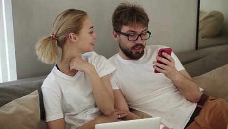 Relaxed-young-couple-watching-smart-phone-together-sitting-on-a-couch-in-a-living-room-at-modern-loft-interior.-Beautiful-young-caucasian-couple-using-phone-at-home.-Man-showing-something-disgusting.-Emotions