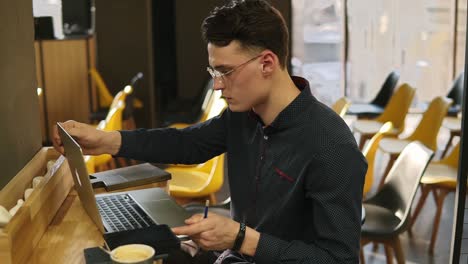 attractive male in his 20's wearing glasses and fitness tracker on his wrist, working in a well lit indoors co working space, typing something on laptop keyboard