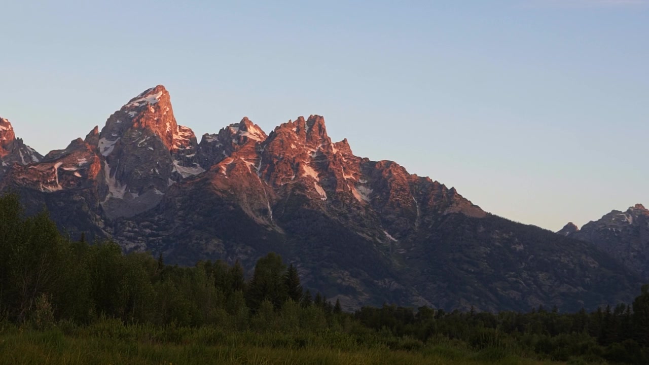 Rekaman gerak lambat close-up yang menyorot ke atas dari pantulan hingga menampakkan matahari terbit menerangi puncak-puncak menjulang Taman Nasional Grand Teton di Schwabacher Landing, dikelilingi semak hijau