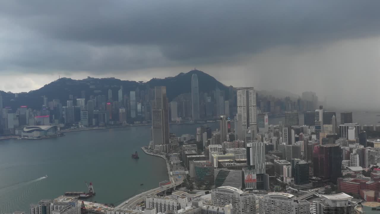 A sweeping aerial view captures Hong Kong's iconic skyline as a dramatic thunderstorm rolls in. The moody, overcast sky contrasts with the vibrant cityscape below