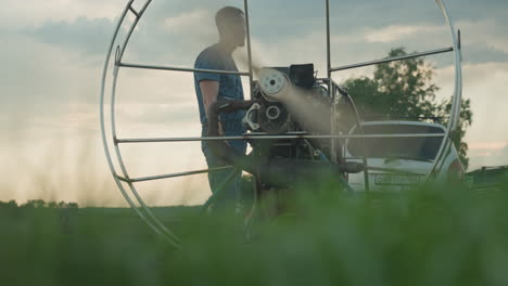 man in blue shirt and gloves stands close to spinning paramotor engine on grassy field at dusk as fan blades whirr within metal cage near vehicle with blurred people in background