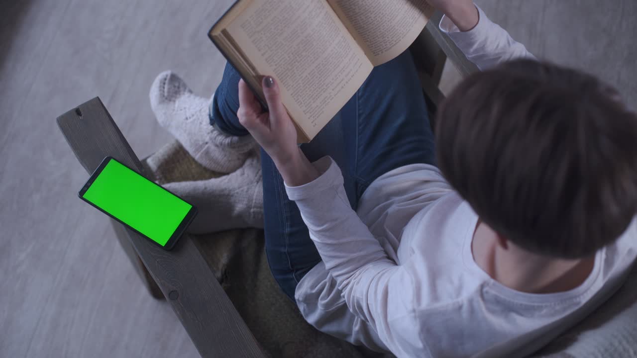Ragazza con i capelli corti in jeans e calzini di lana seduta in poltrona a casa che legge un libro in una mattina d'inverno. Accanto un telefono cellulare con schermo verde. Concetto di nuova tecnologia: cellulare con schermo verde e chroma key.