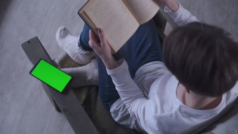 niña con cabello corto en vaqueros y calcetines de lana se sienta en casa en un sillón y lee un libro en una mañana de invierno. cerca de un teléfono móvil pantalla verde. clave de croma teléfono celular pantalla verde nuevo concepto de tecnología.