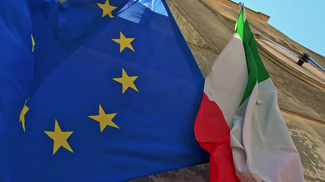 Close-up view of European and Italian flags fluttering together against a stone building under a clear sky.