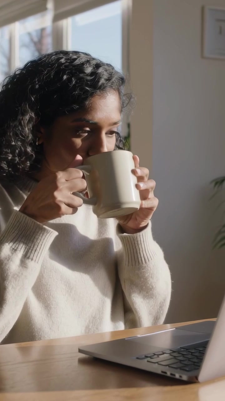 Video vertical: Mujer con suéter ligero pausando la escritura, alcanzando una taza para beber en casa, con el portátil abierto.