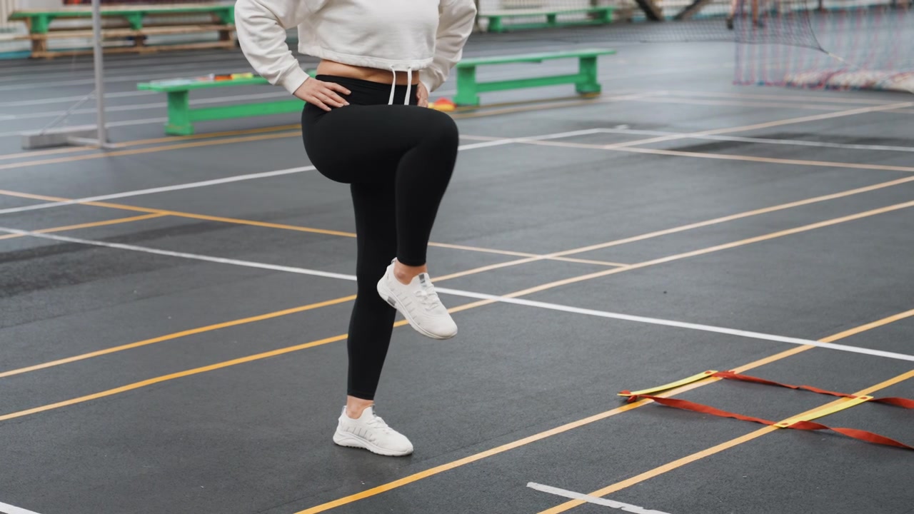 Secuencia de agilidad con escalera de calentamiento en cancha exterior, con práctica de juego de pies inspirada en el bádminton. Atleta solitaria concentrada realizando elevaciones de rodilla, saltos laterales y ejercicios de equilibrio en una superficie marcada.