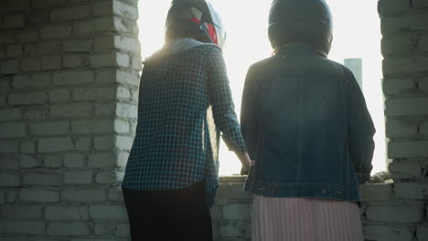 back view of two women standing at a window, one wearing black leggings and sneakers, the other in a pleated skirt and denim jacket, both appear calm