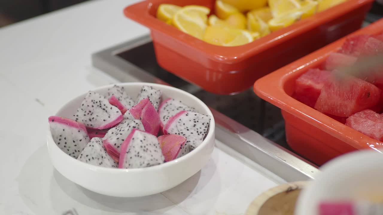A close-up of a vibrant fruit buffet featuring dragon fruit, watermelon, and citrus, with a hand using tongs to select fruit