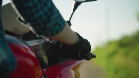 primer plano de la mano de un motociclista con guante y camisa a cuadros, acelerando una motocicleta, el fondo está borroso con un entorno verde natural