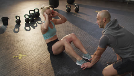 Fit-caucasian-woman-performing-crunches-while-male-trainer-holding-her-legs-at-the-gym