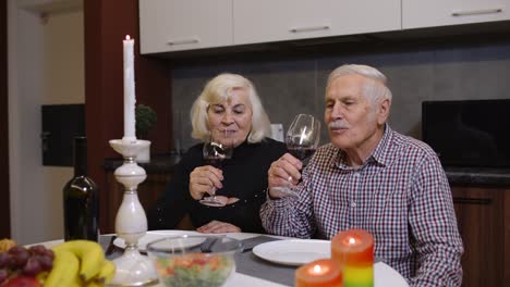 Portrait-of-senior-old-couple-toasting-wine-at-home-during-romantic-supper-in-kitchen-at-home