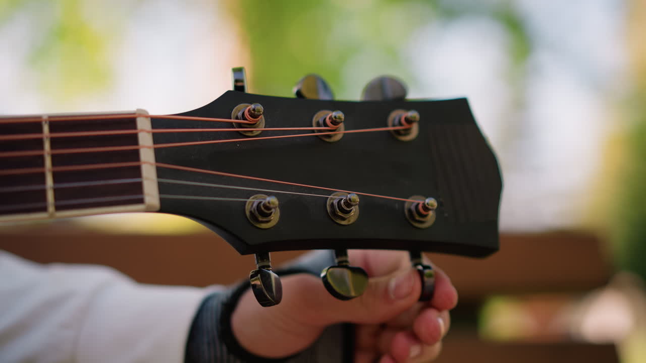white hand adjusting guitar tuning peg outdoors in park, closeup headstock and fretboard, sunlight filtering through leaves, glovecovered wrist, musician prepping for gig, focus on precision
