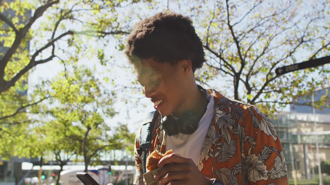 Happy african american man in city, using smartphone, wearing headphones, holding pastry in street