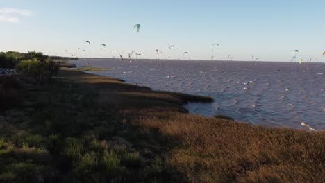 aerial forward at low altitude over vicente lopez park with many kitesurfers on rio de la plata river at sunset, buenos aires in argentina