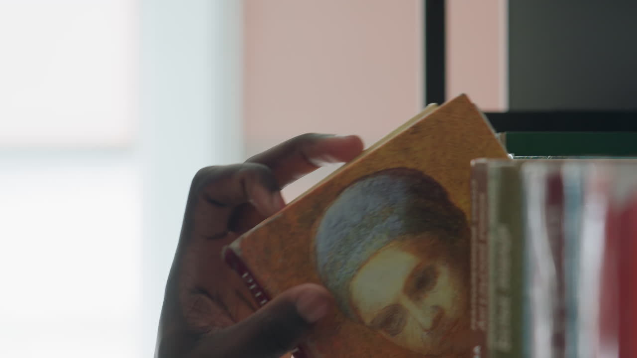 Close up of dark skin hand reaching to pick book from shelf, fingers gently touching spine with blurred background, capturing detail of natural light, and quiet focused atmosphere indoors