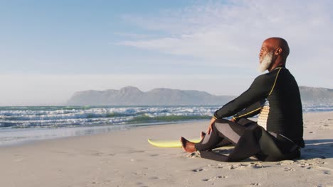 hombre afroamericano mayor sentado con una tabla de surf en la playa